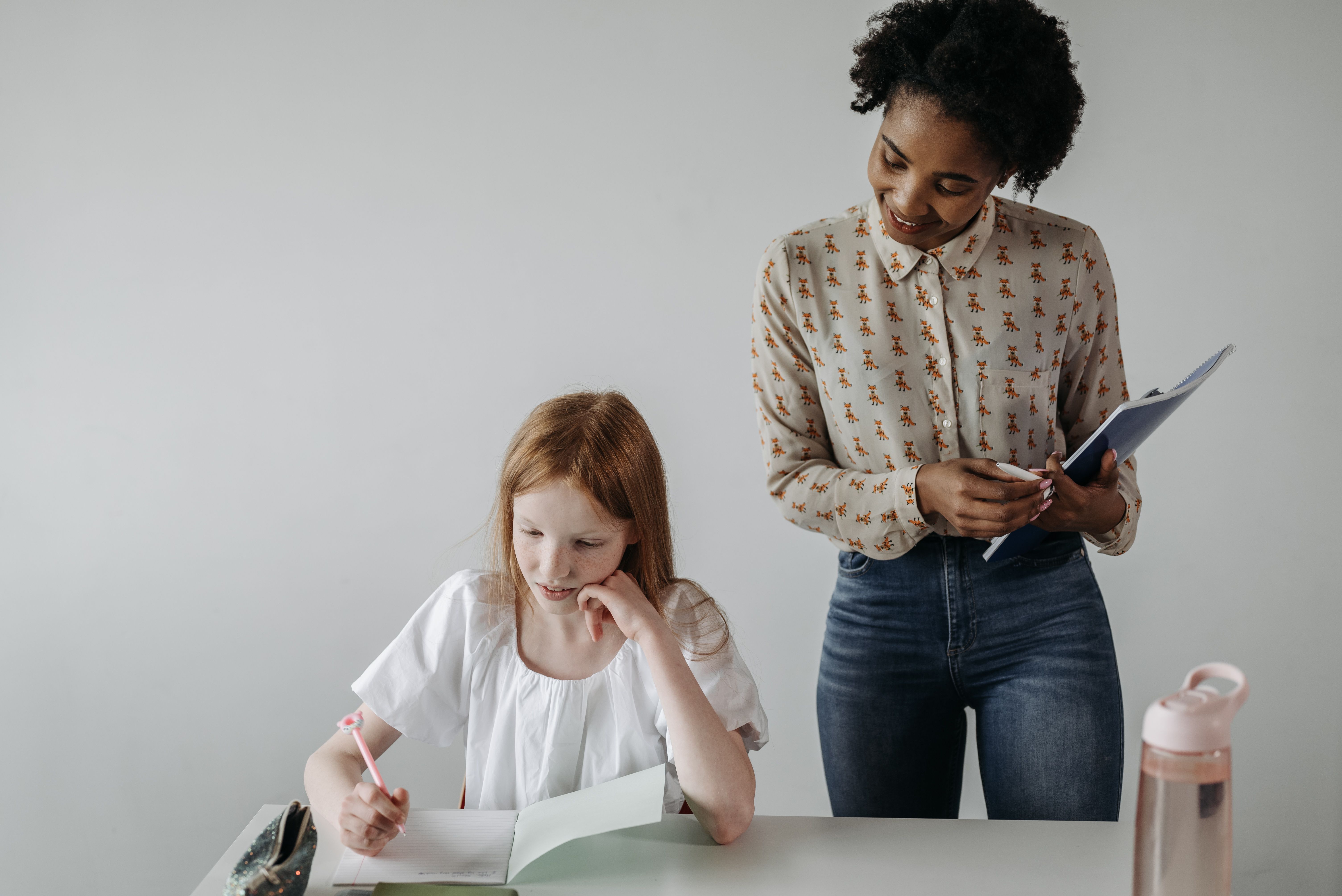 A teacher guides a student who is writing at a classroom desk